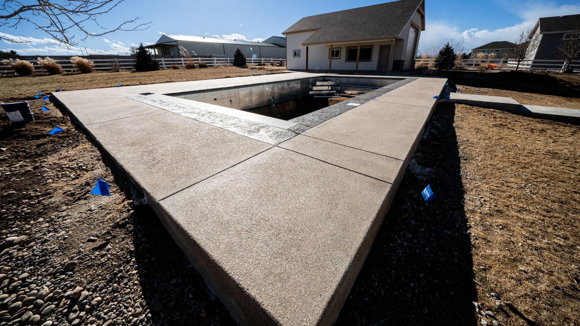 Concrete courtyard walkway surrounding a rectangular pool under construction, beside a house and gravel yard.