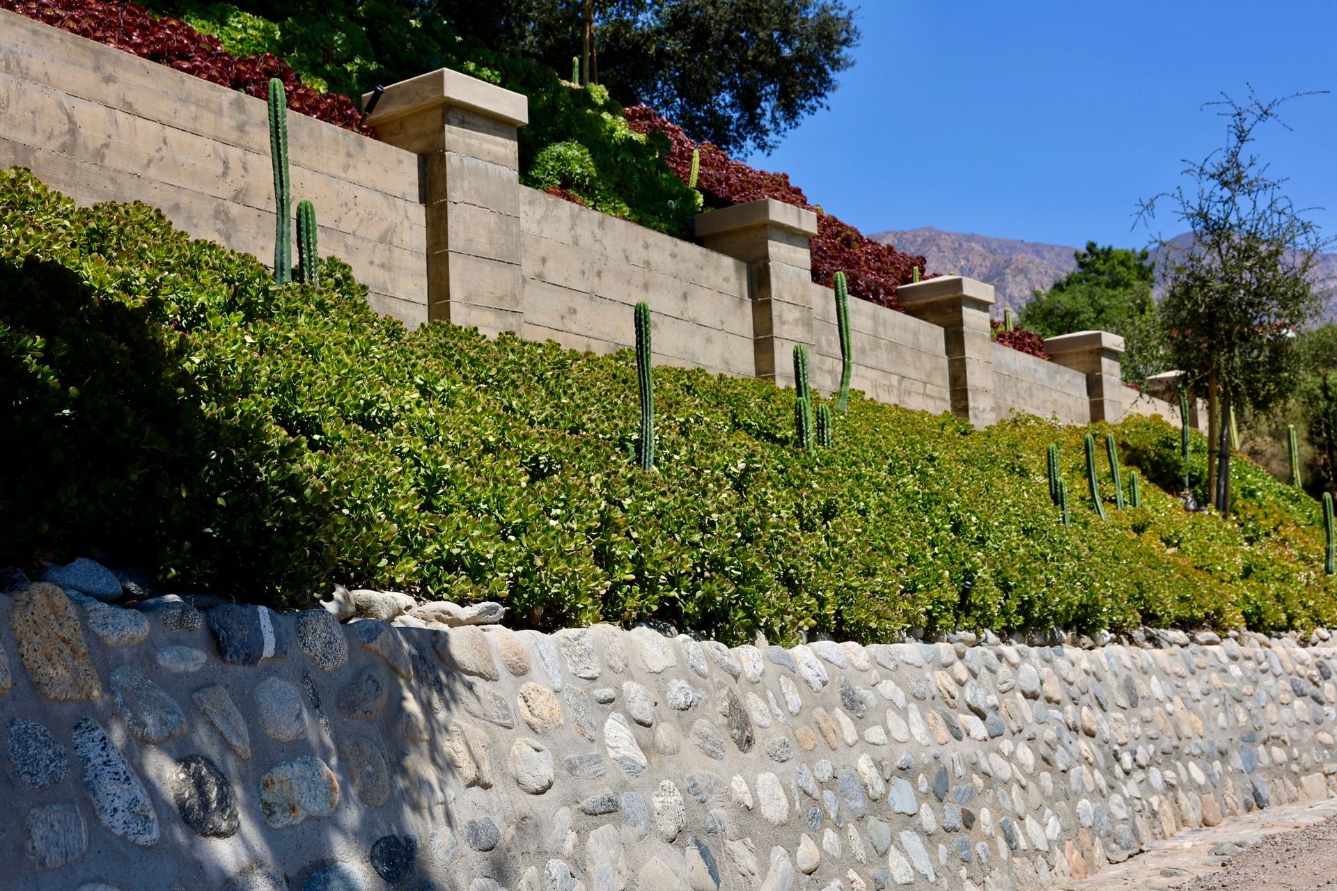 Stone retaining wall below a lush green hillside with tan fence panels and trees under a blue sky