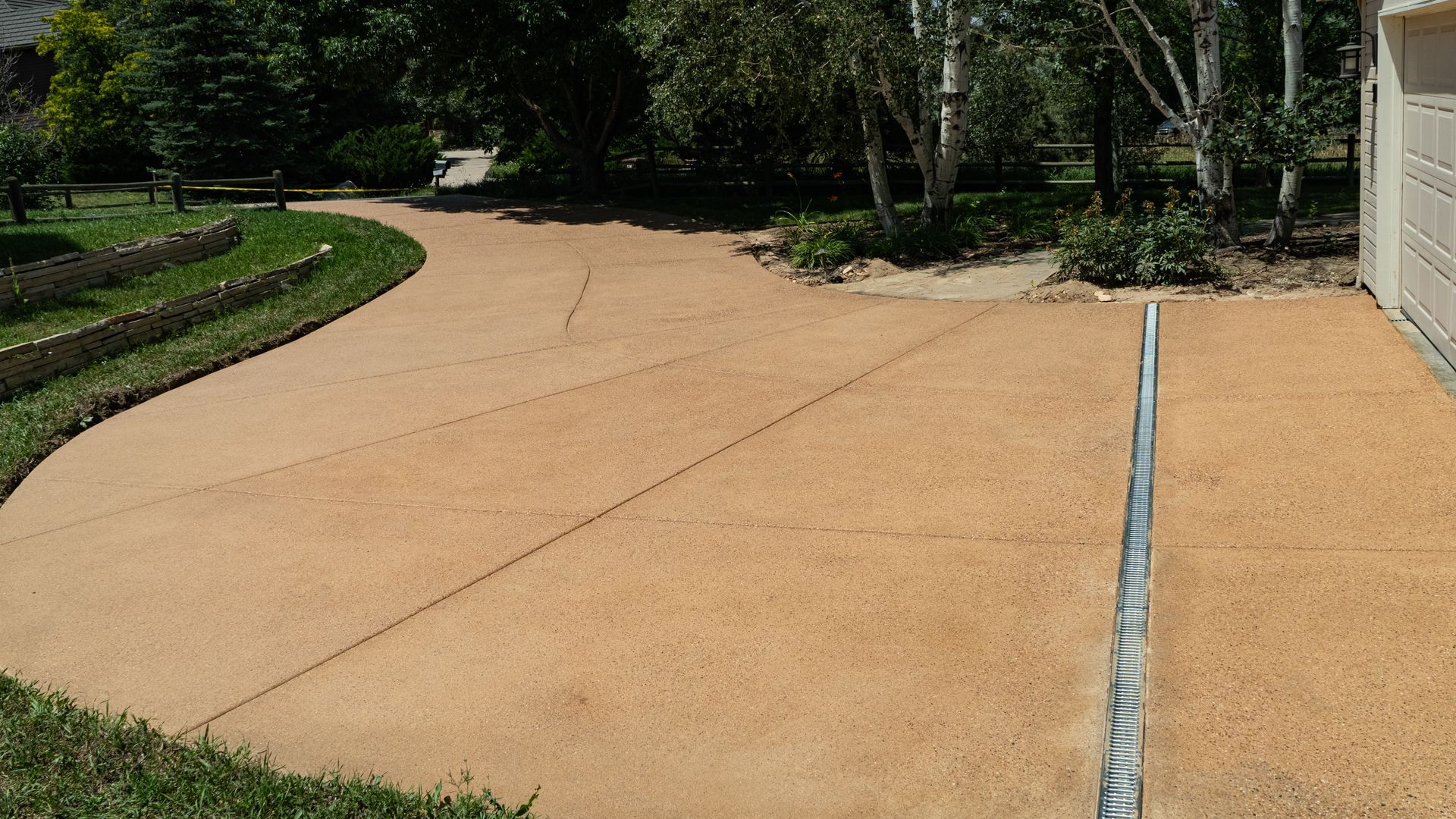 Tan concrete driveway curving beside a green garden and trees next to a house