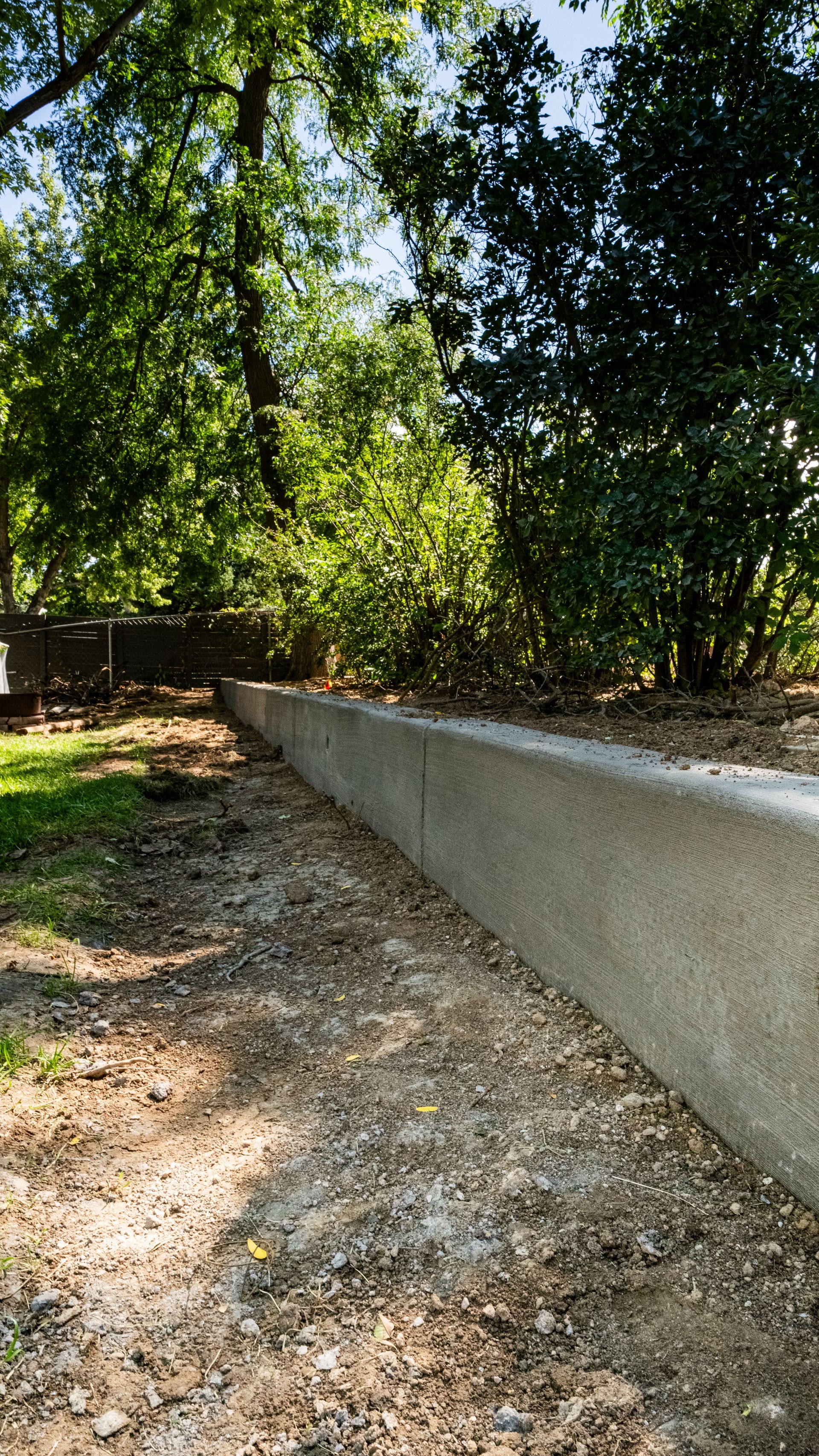 Gravel path beside a white retaining wall in a wooded area with sunlight filtering through trees