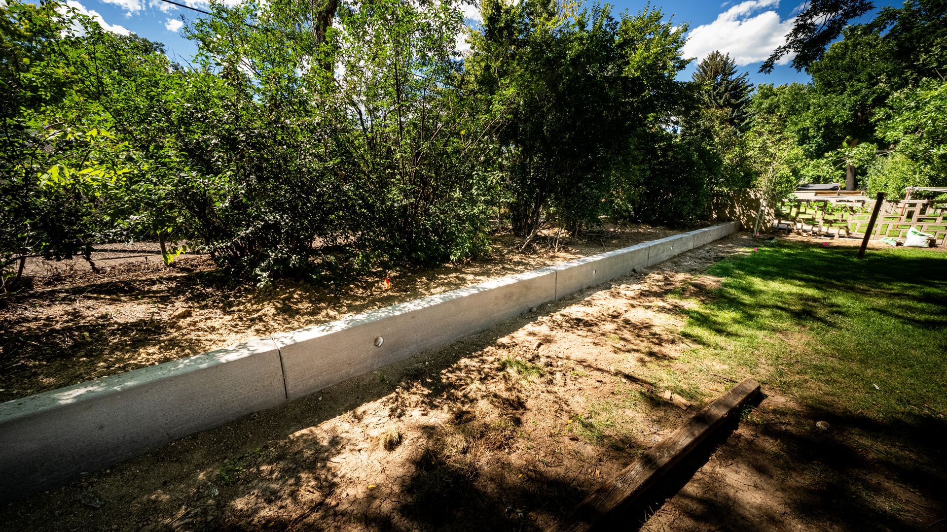 Concrete garden border along a dirt path beside green trees and shrubs under a sunny sky.