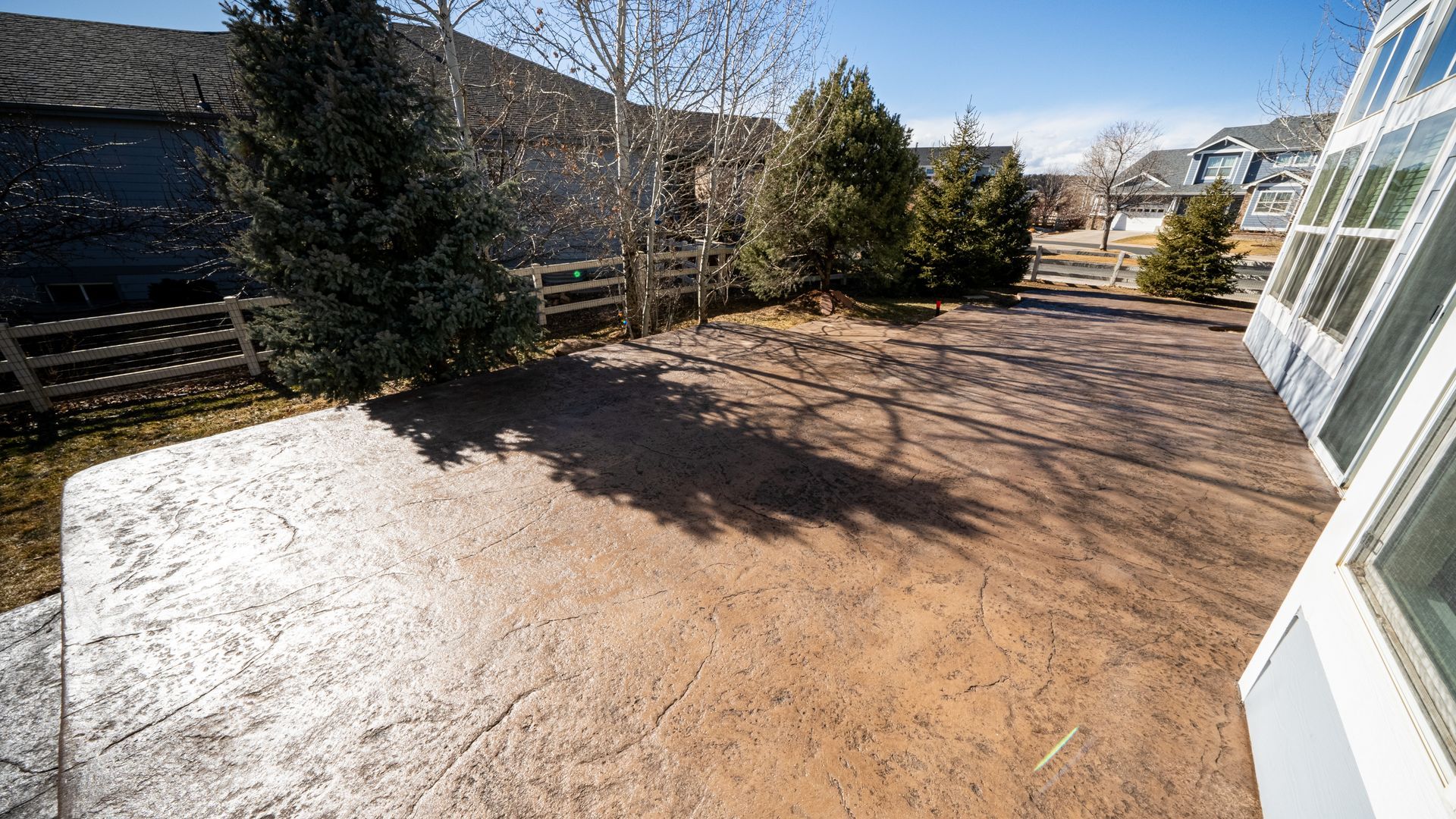 Sunny backyard patio with a large concrete surface, shrubs, and glass doors on the right.
