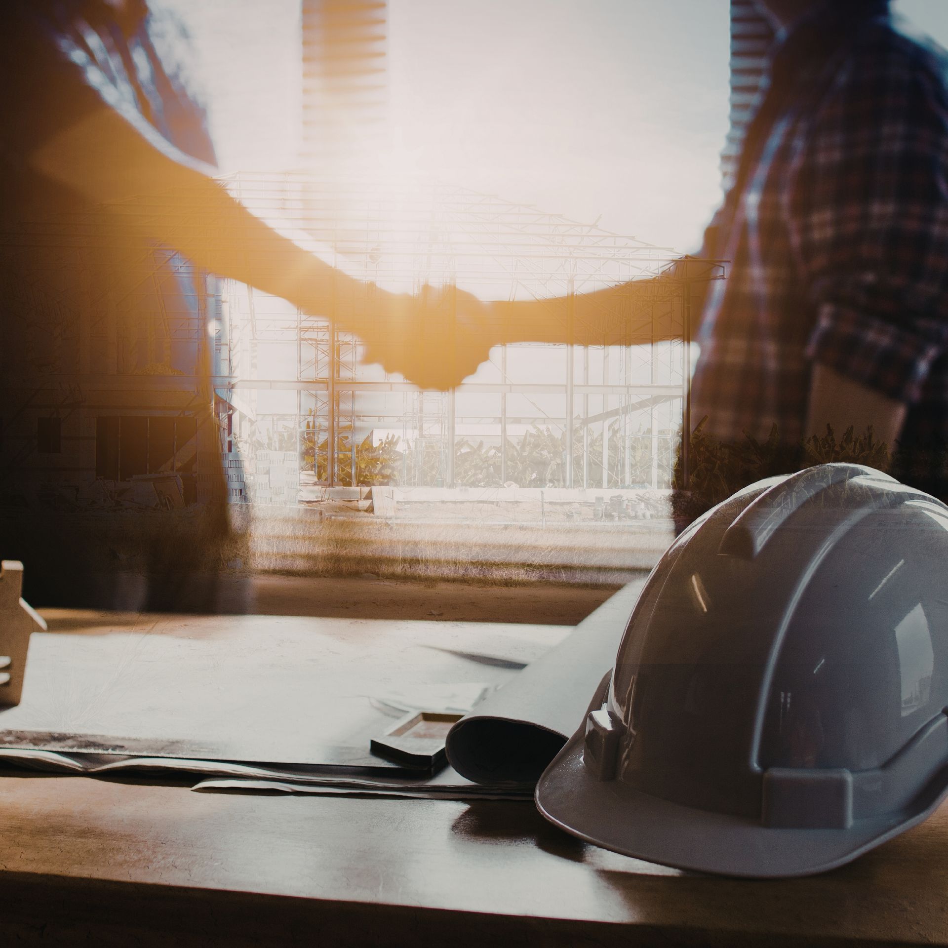 Two people shake hands at a construction site desk with a hard hat and plans in warm sunlight