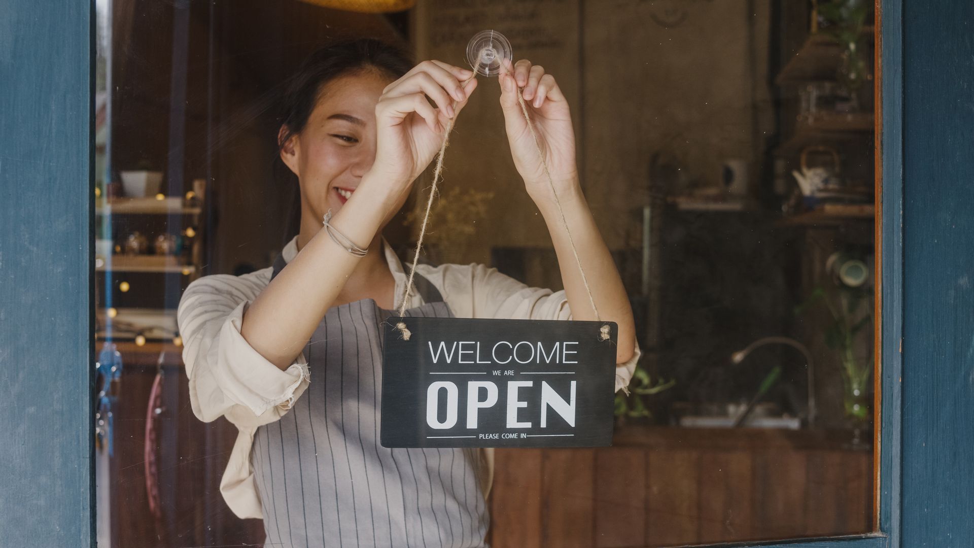 Shop owner hanging a “WELCOME OPEN” sign in a cozy storefront window.