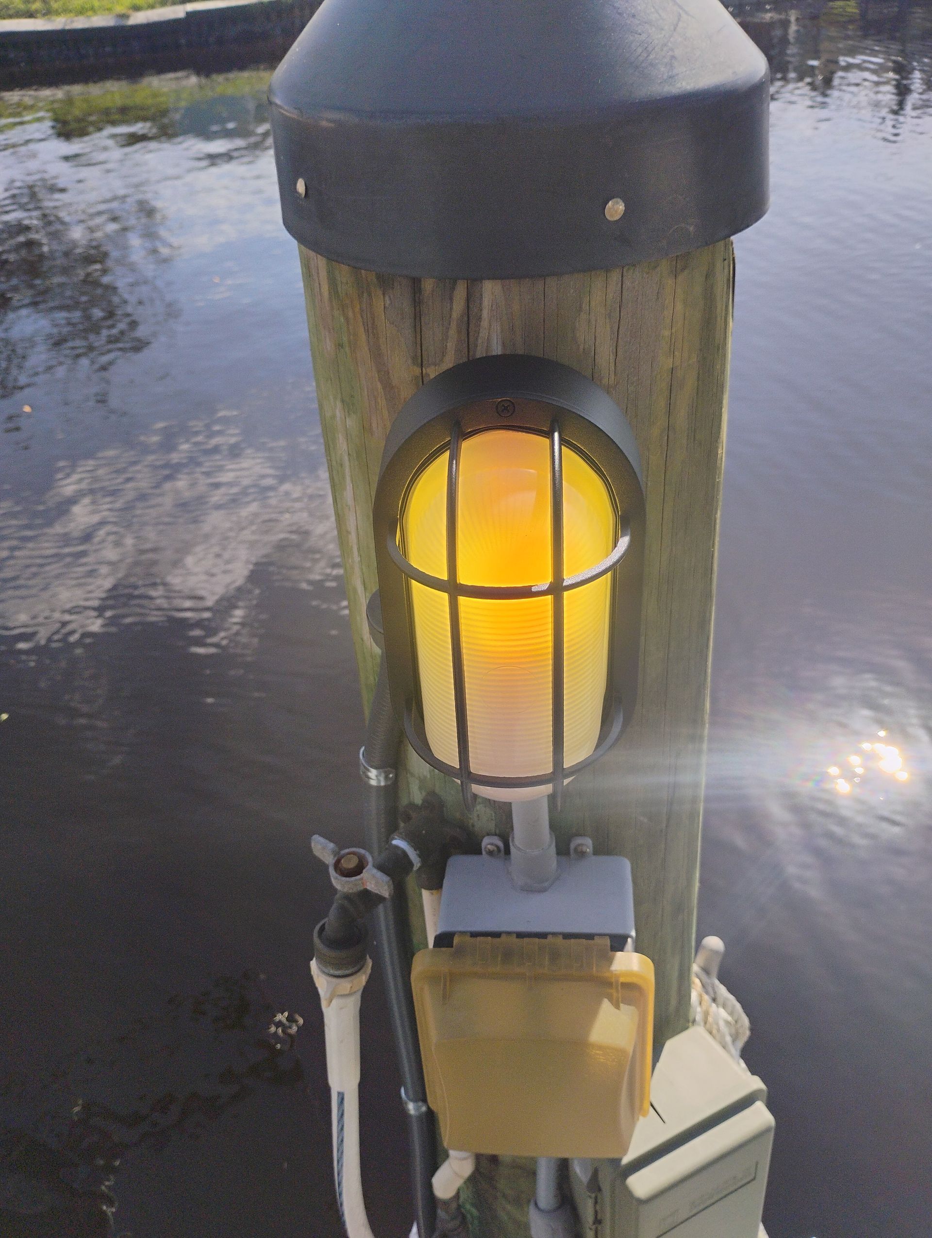 Yellow-lit security lamp on a wooden dock piling near water. Black metal cage protects the light.