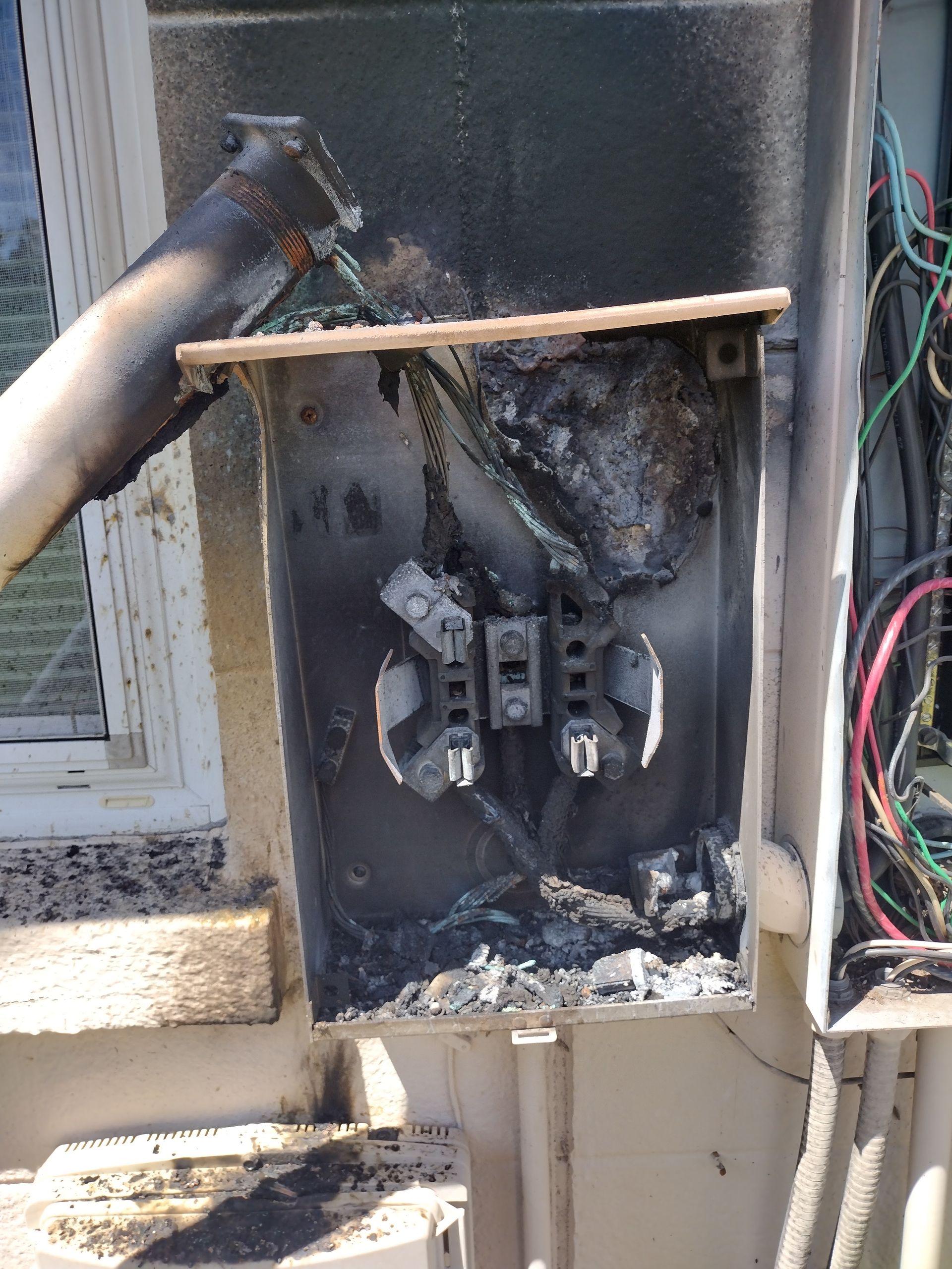 Burned-out electrical meter box, charred interior. Blackened wires, melted components, near a window.