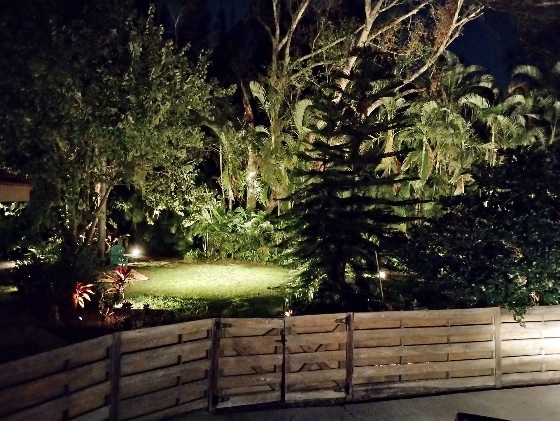 Nighttime view of a lit backyard with a wooden fence in the foreground and trees, plants, and a small lawn.