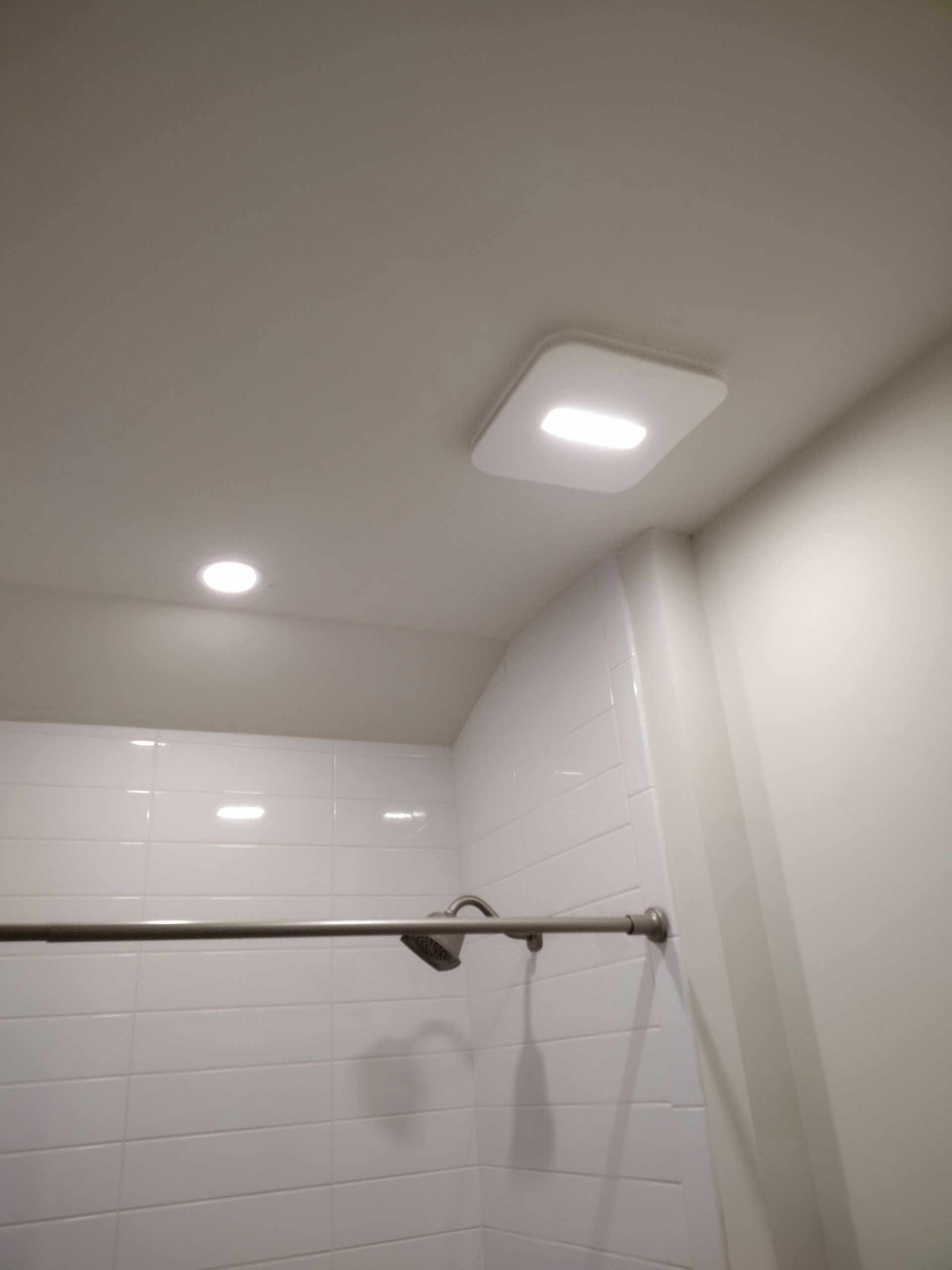 Shower interior with white subway tile, showerhead, and ceiling lights, including a square ventilation fan.