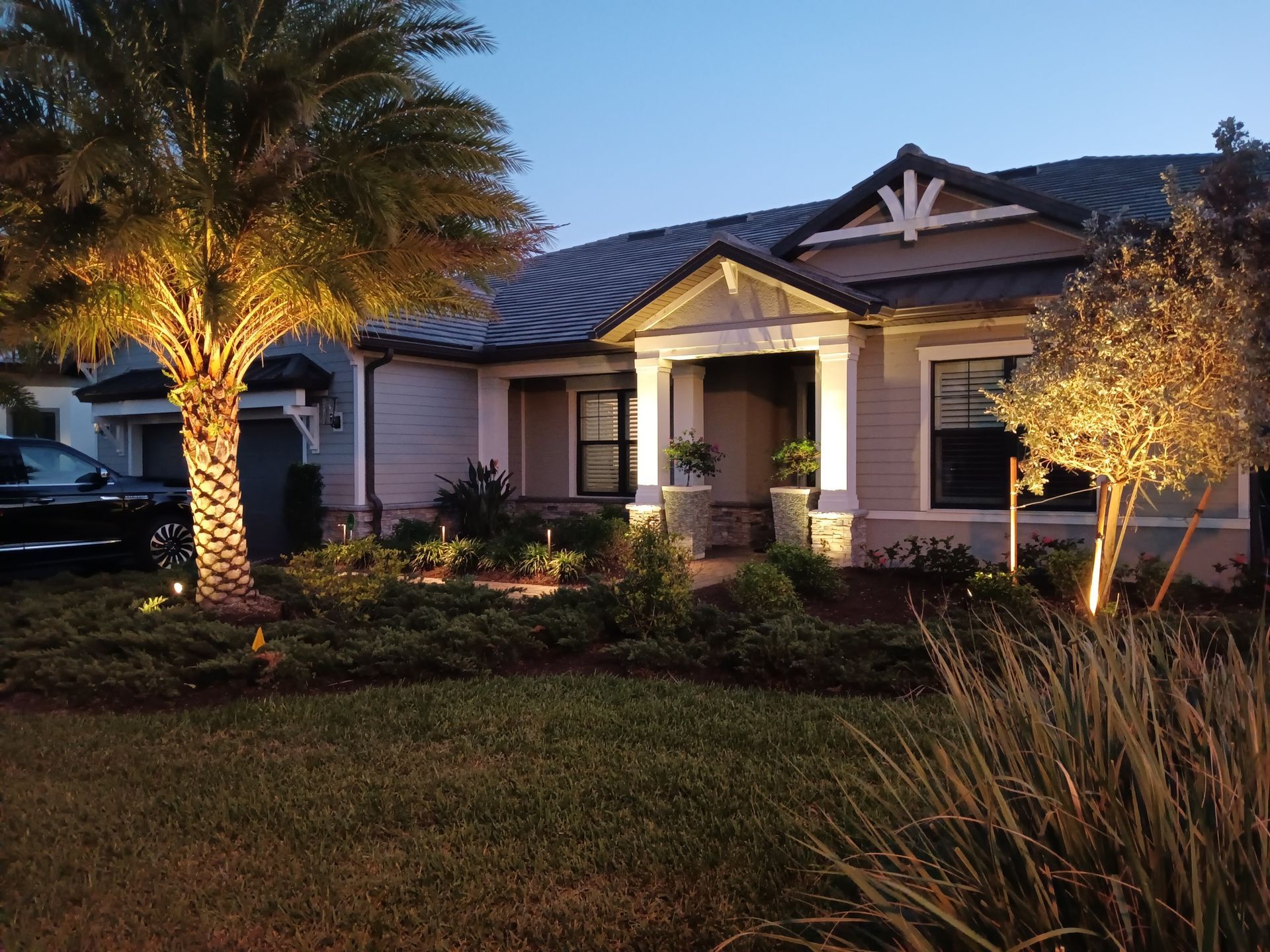 House exterior at dusk, illuminated landscaping. Palm tree on left, shrubbery, and a black car.