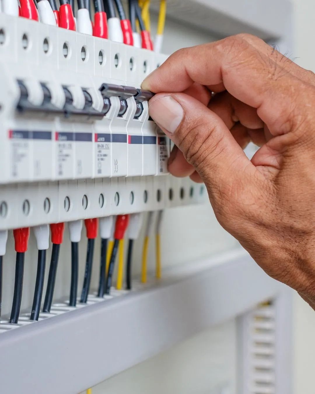 Person's hand flips a circuit breaker switch in an electrical panel, with red and black wires visible.