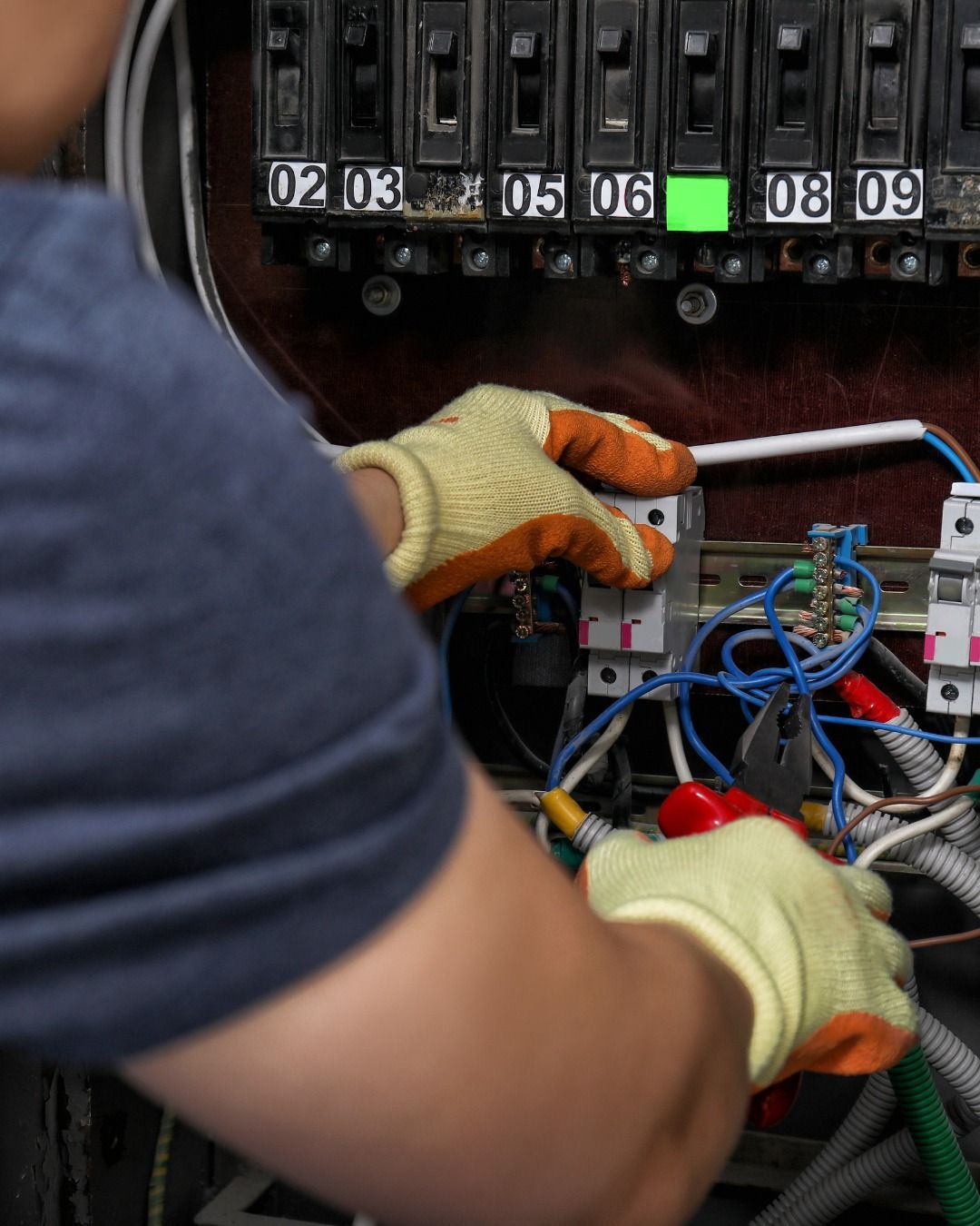 Electrician working on an electrical panel, wearing gloves, with various wires and circuit breakers.