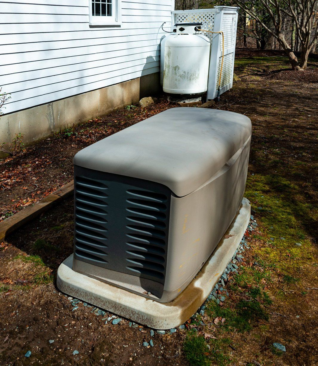 Generator with a beige cover on a concrete base, next to a white propane tank near a house.