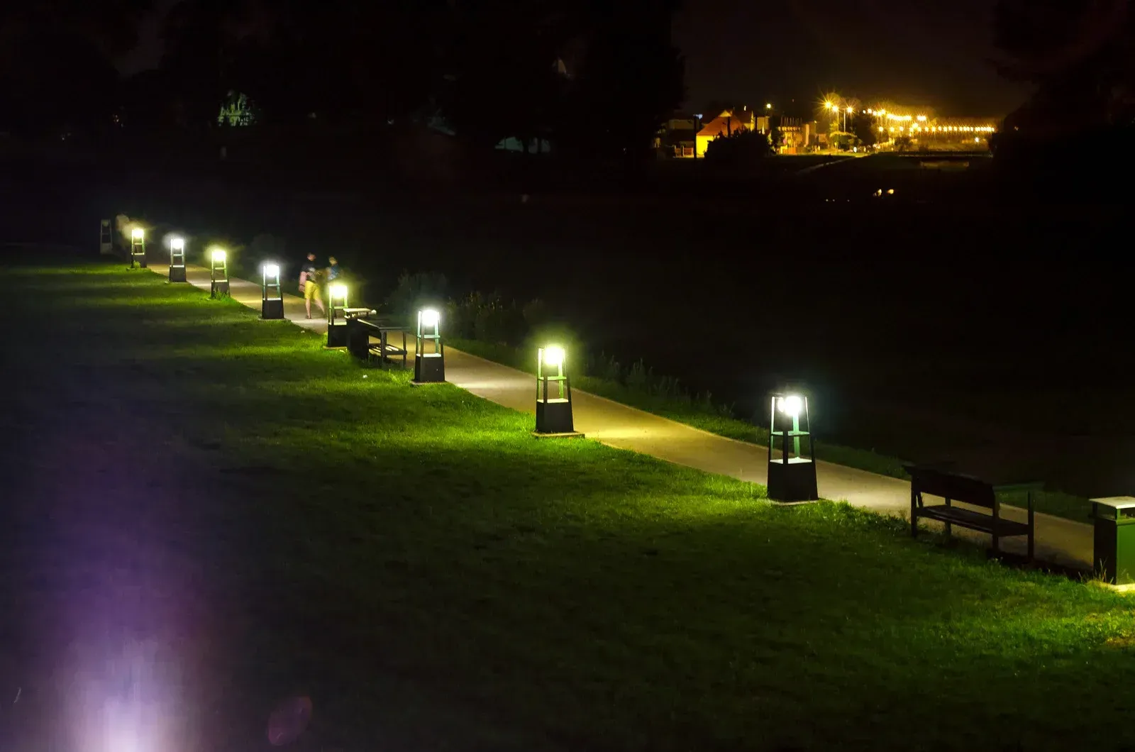 Nighttime view of a path lined with illuminated lampposts, green grass, and benches. A distant cityscape glows.