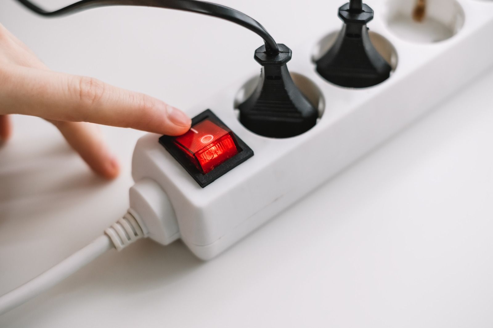 A hand pressing a red power switch on a white power strip with black plugs inserted.