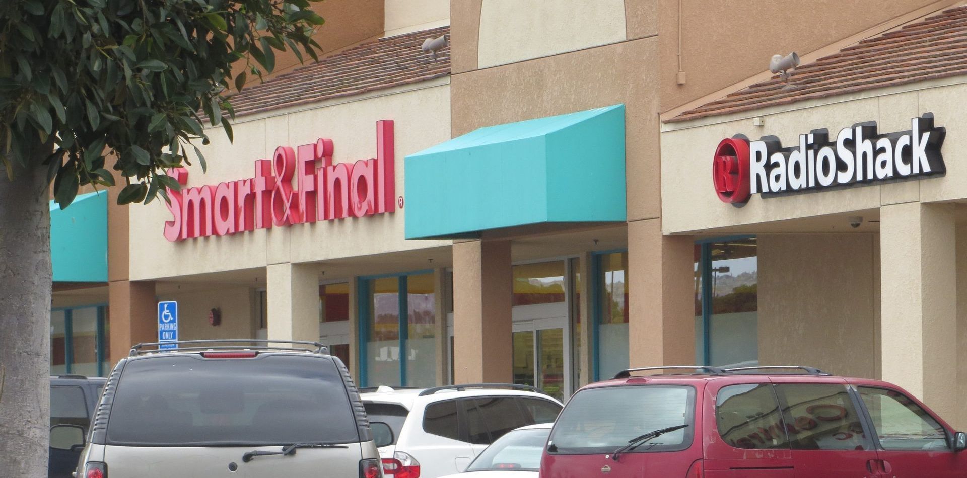 A red car is parked in front of a store called radio shack and a light teal awning