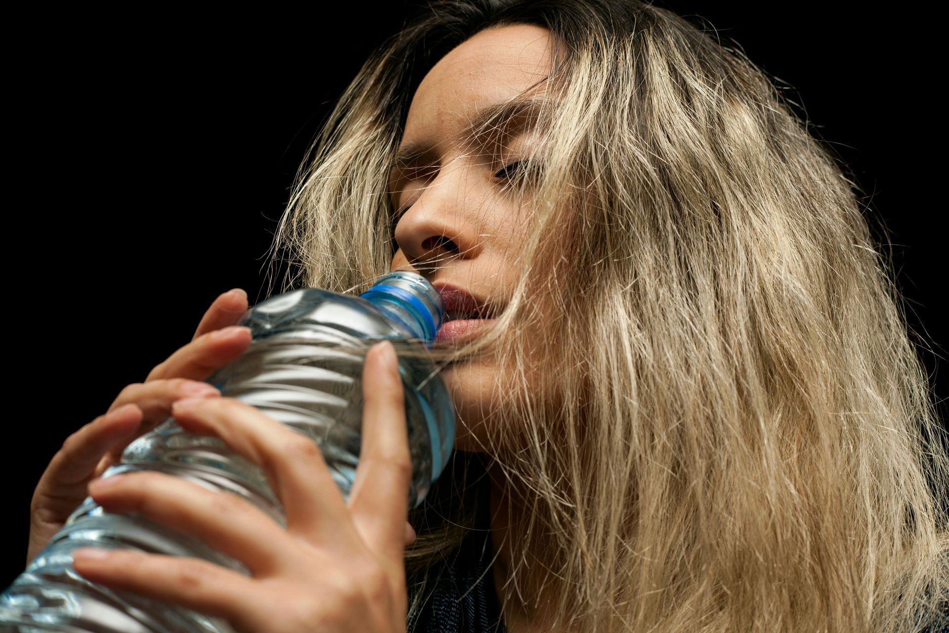 Woman with blonde hair drinks from a clear plastic water bottle, eyes closed