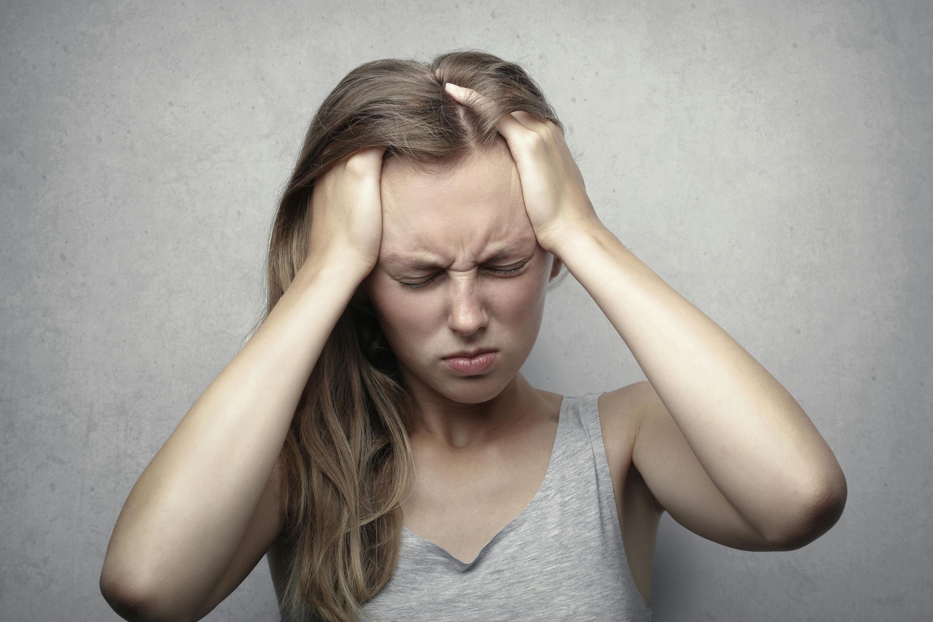 Woman holding head, eyes squeezed shut, in distress; gray tank top.
