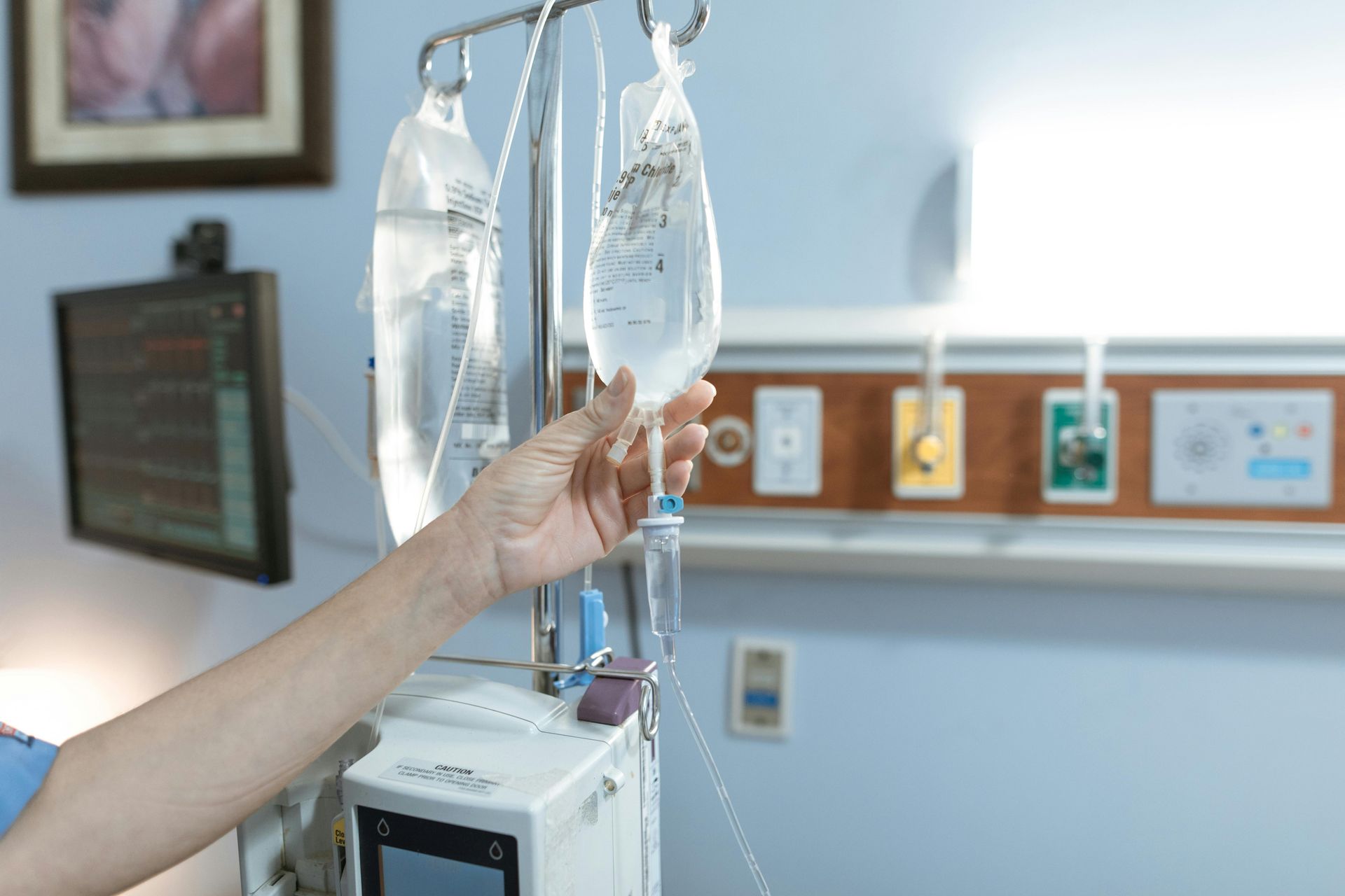Hand adjusting IV bag in a hospital room with medical equipment and blue walls.