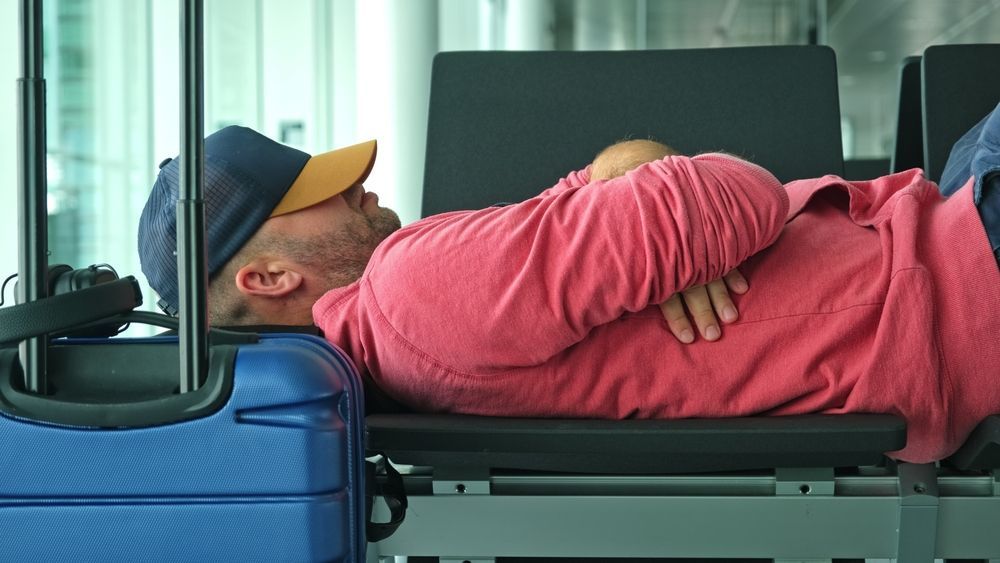 A man is sleeping on a bench next to a blue suitcase.