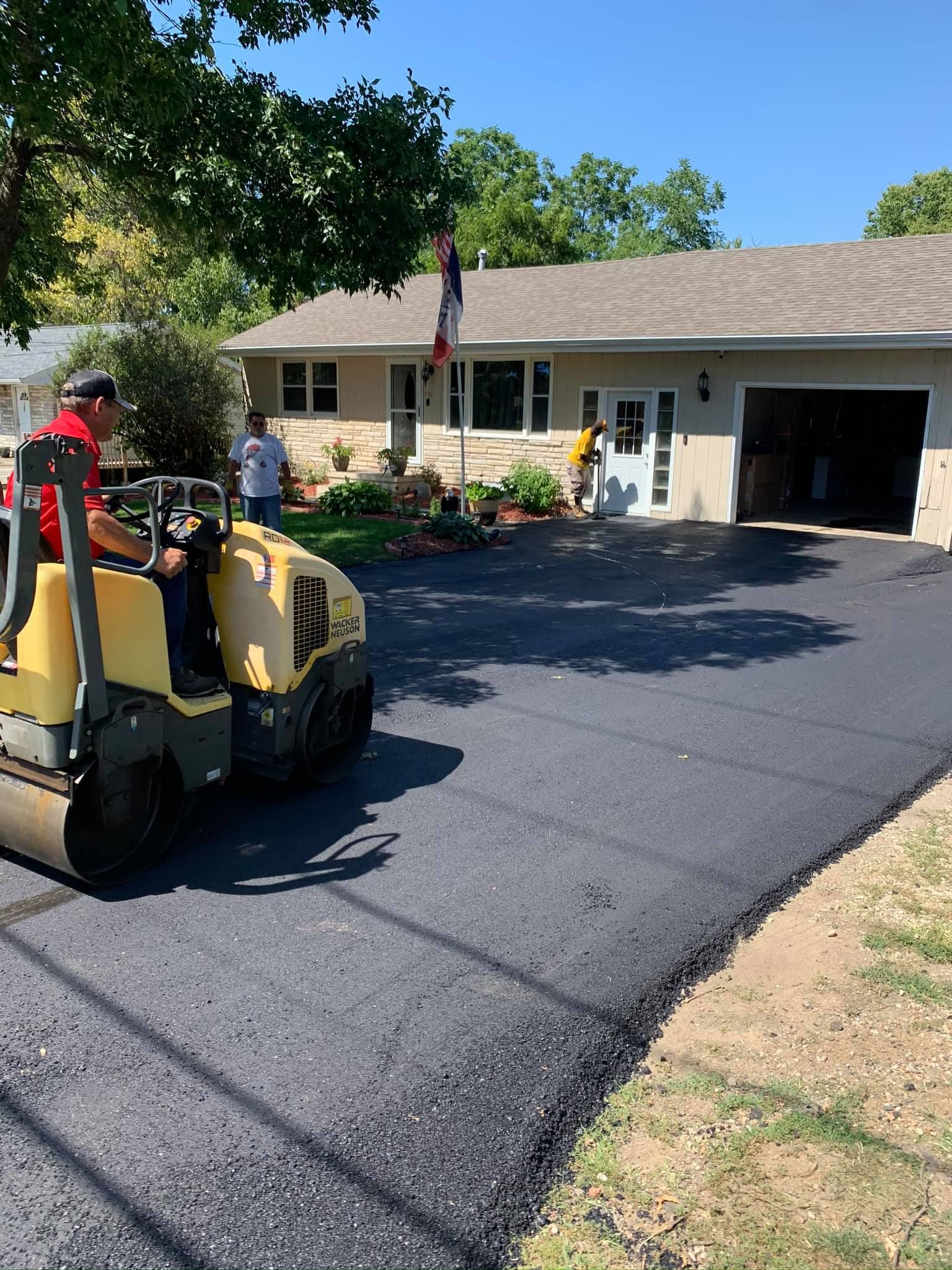 Worker operating a road roller, paving a driveway in front of a house on a sunny day.
