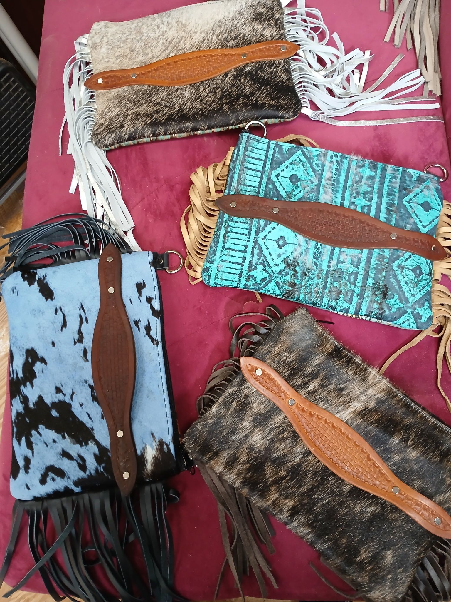 Cowhide purses in blue, turquoise, and black with leather accents and fringe on a red surface.
