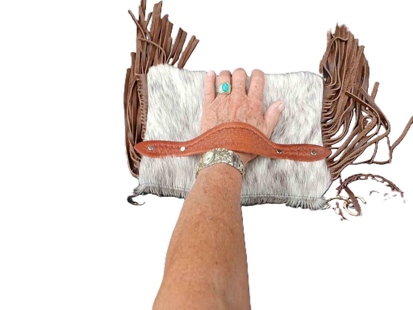 Hand holding a white and gray fur clutch with leather strap and fringe.