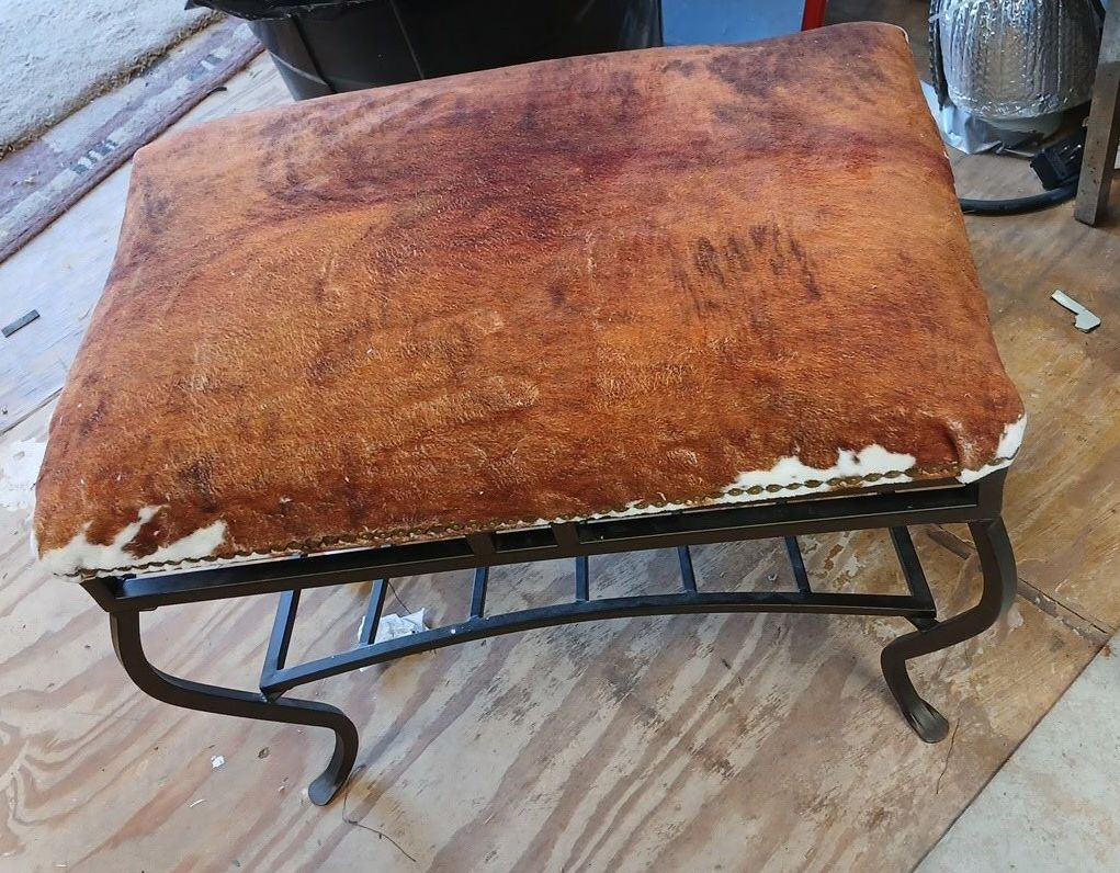 Worn brown ottoman with a rusted metal base on a wood surface. The cushion is heavily stained.