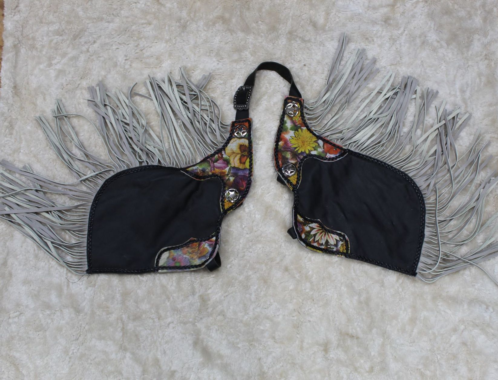 Black chaps with colorful floral designs, white fringe, and an adjustable connecting strap, resting on a white, textured surface.