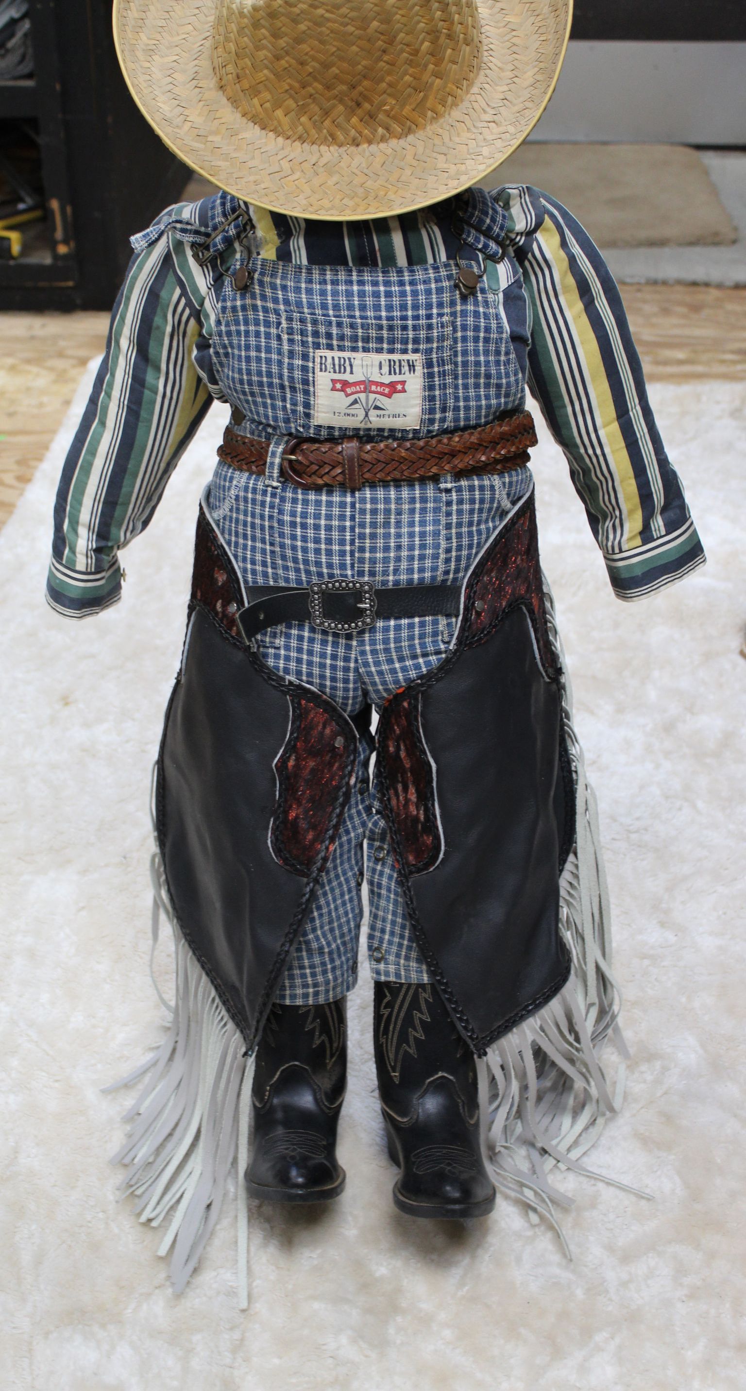 Doll dressed as a cowboy. It wears a plaid shirt, chaps, belt, boots, and a straw hat.