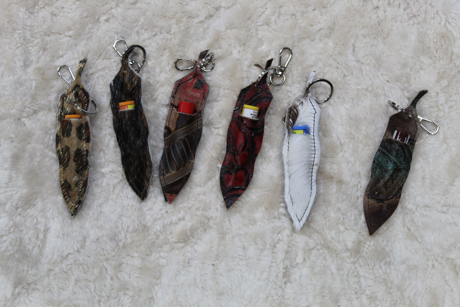 Six leather-textured keychains in varying colors and textures, arranged on a textured, light-colored surface.