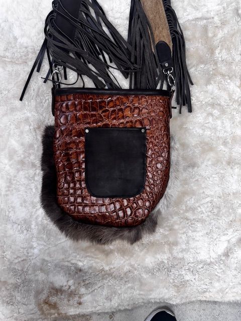 Brown leather crossbody bag with fringe, alligator-like texture, and a black pocket on a fur background.