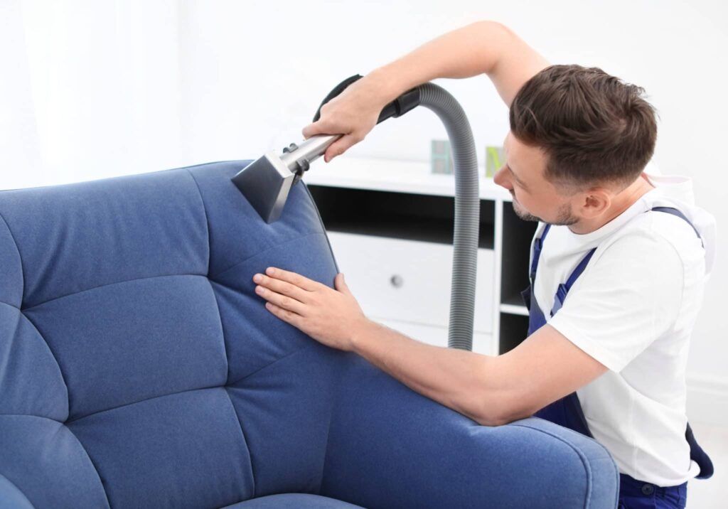 A man is cleaning a blue couch with a vacuum cleaner.