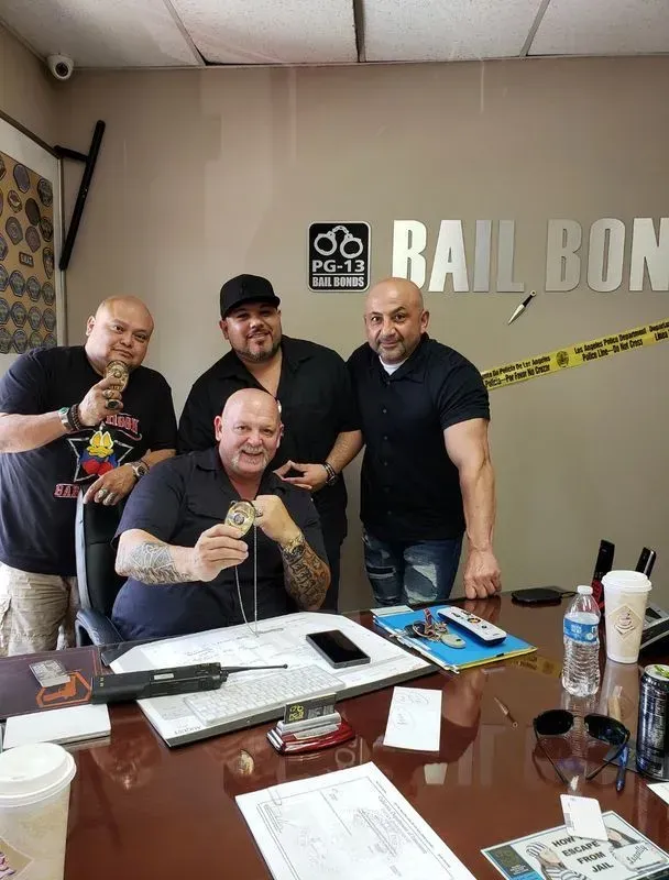 Four men pose in a office, one seated at a desk holding a trophy, with a “Bail Bond” sign behind them. Four men pose in a office, one seated at a desk holding a trophy, with a “Bail Bond” sign behind them.