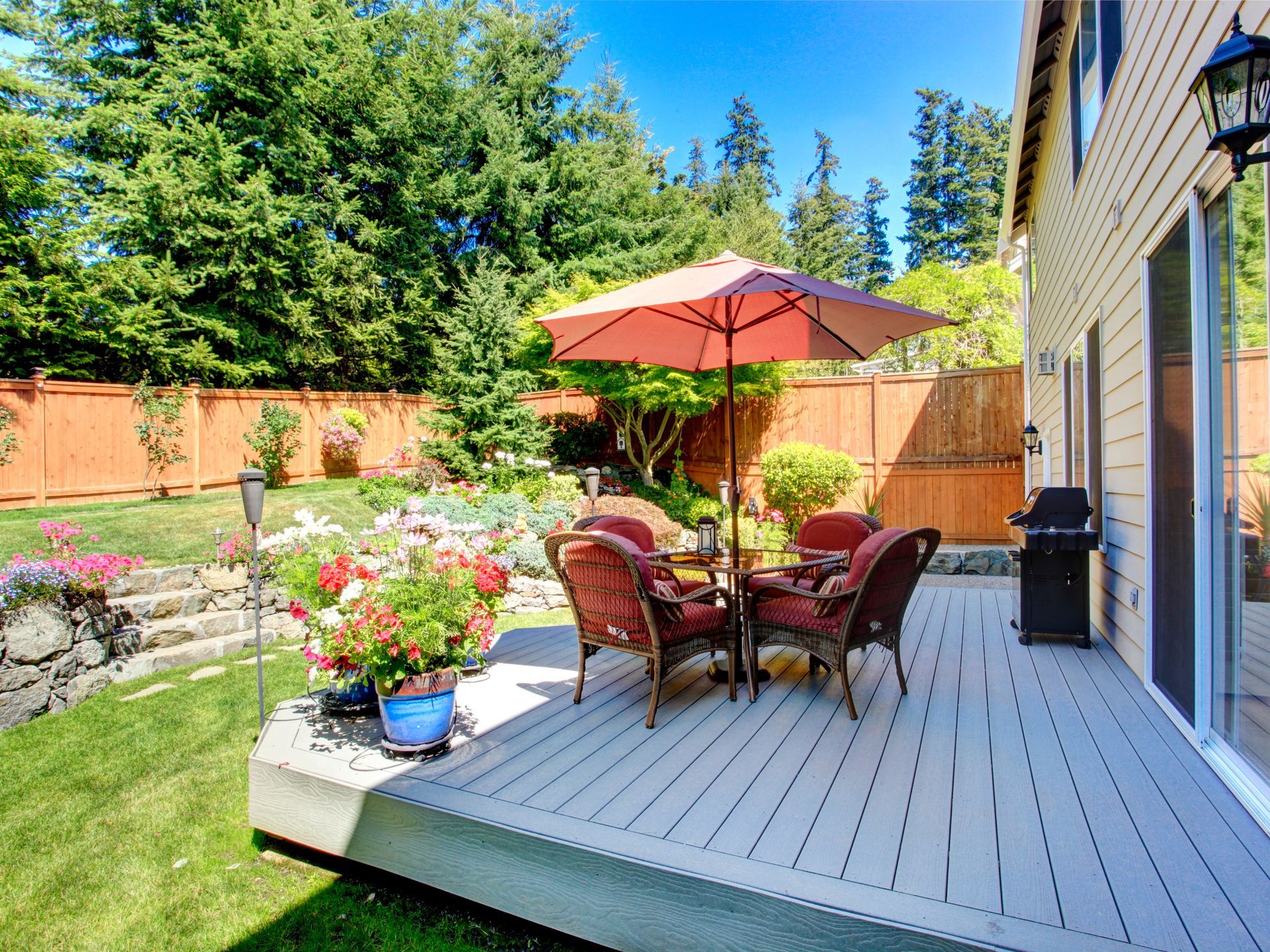 Backyard deck with table, chairs, and umbrella, surrounded by greenery, flowers, and a wooden fence.
