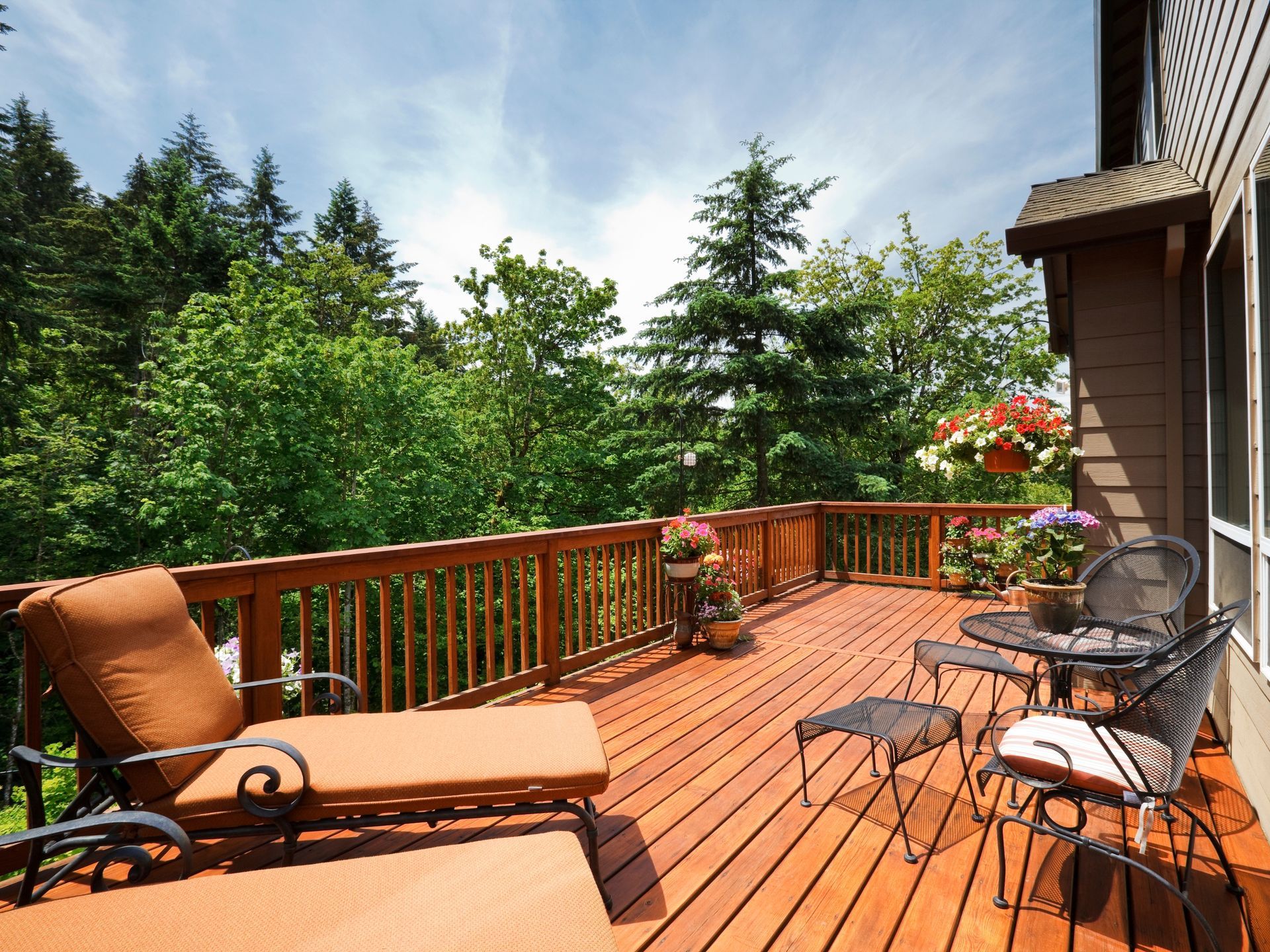 Wooden deck with lounge chair and small table, overlooking lush green trees under a blue sky.