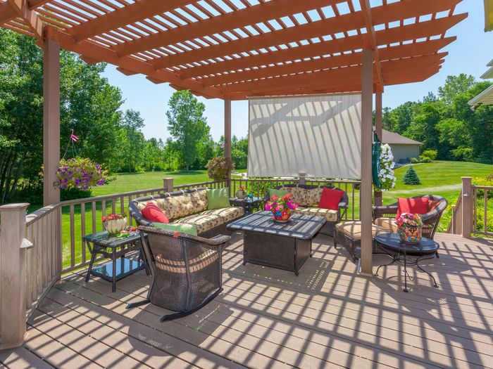 Outdoor deck with seating under a pergola, overlooking a green lawn. Brown wooden structure, colorful cushions.