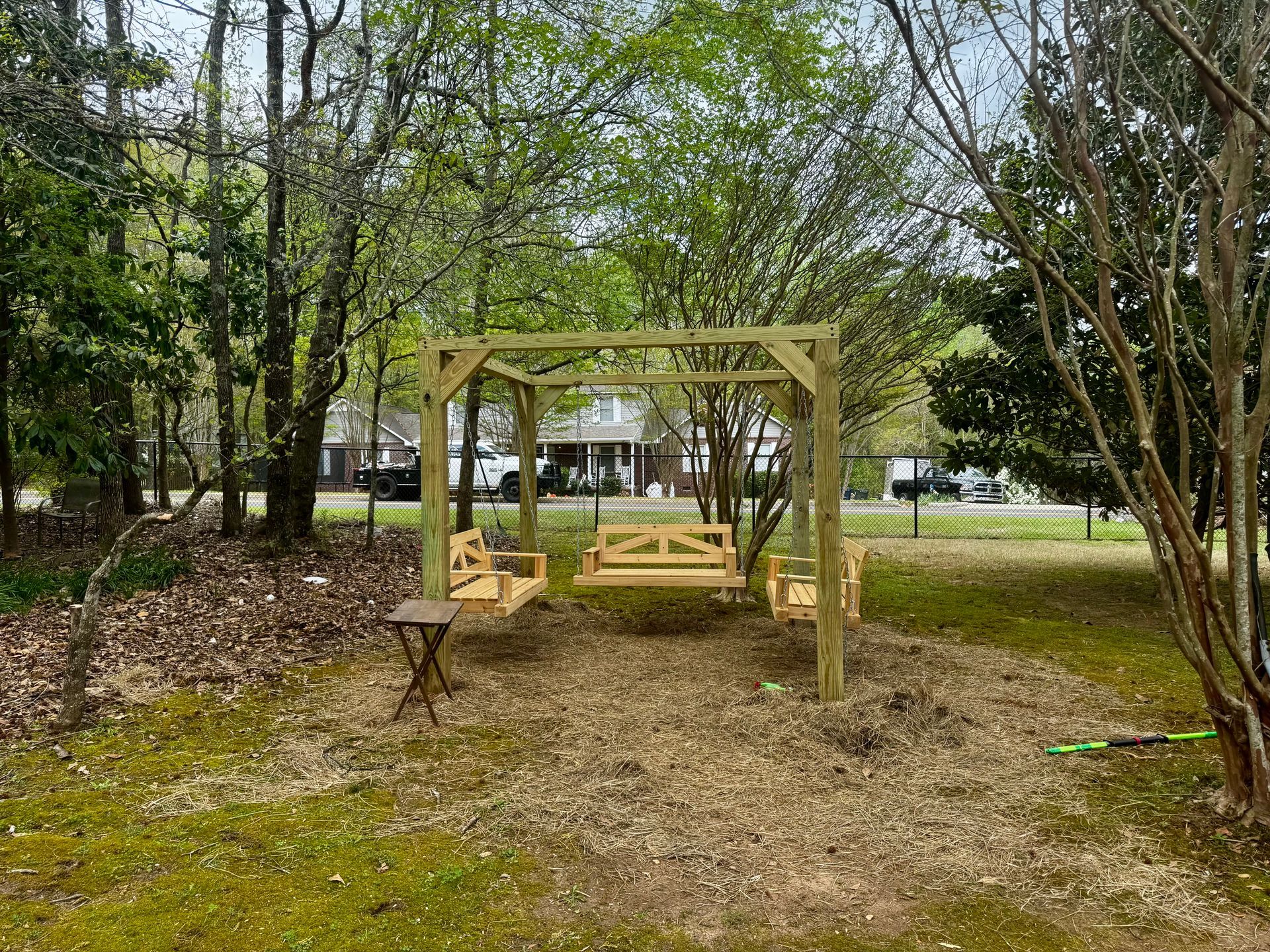 Wooden swing set in a yard, surrounded by trees and mulch. A small table is nearby.