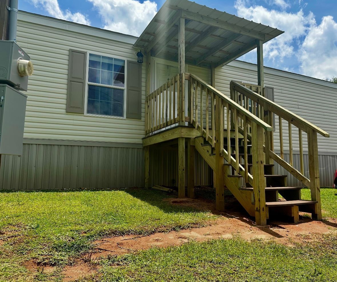 Mobile home with wooden porch and stairs, surrounded by grass, under a sunny sky.