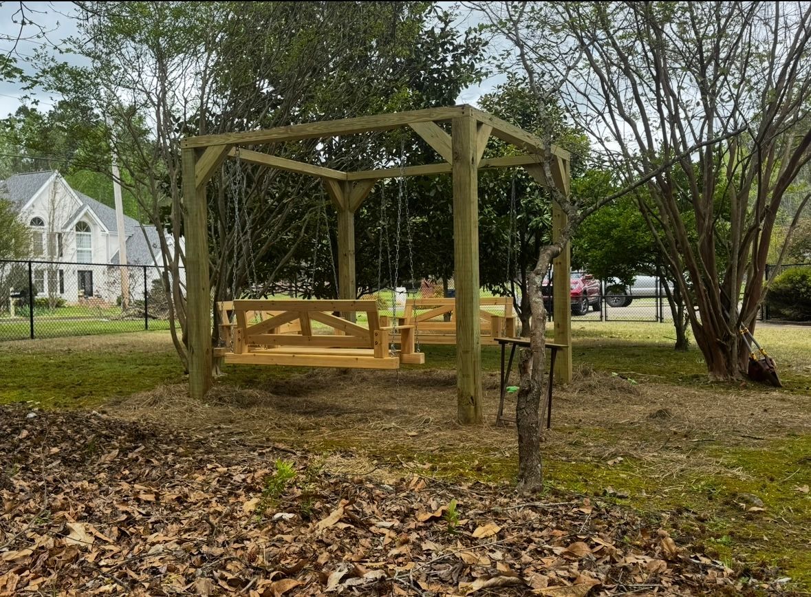 Wooden swing set in a yard, featuring swings and a gazebo-like frame. Green trees and a house in the background.
