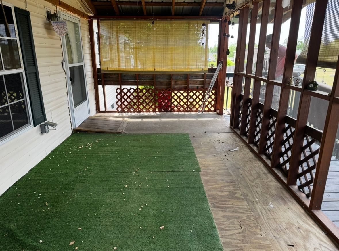 Covered porch with green turf rug and exposed wooden floorboards, latticed railing.