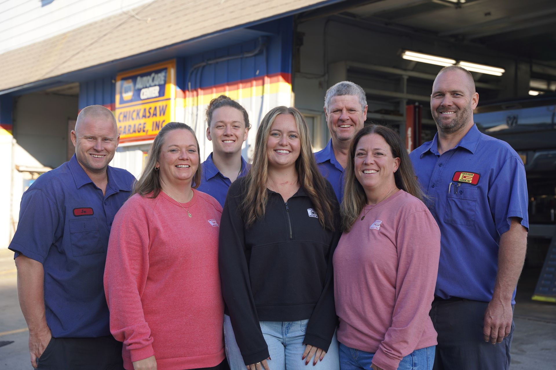 Group of seven people smiling in front of a garage. Building has a blue exterior with a Chickasaw Garage sign.