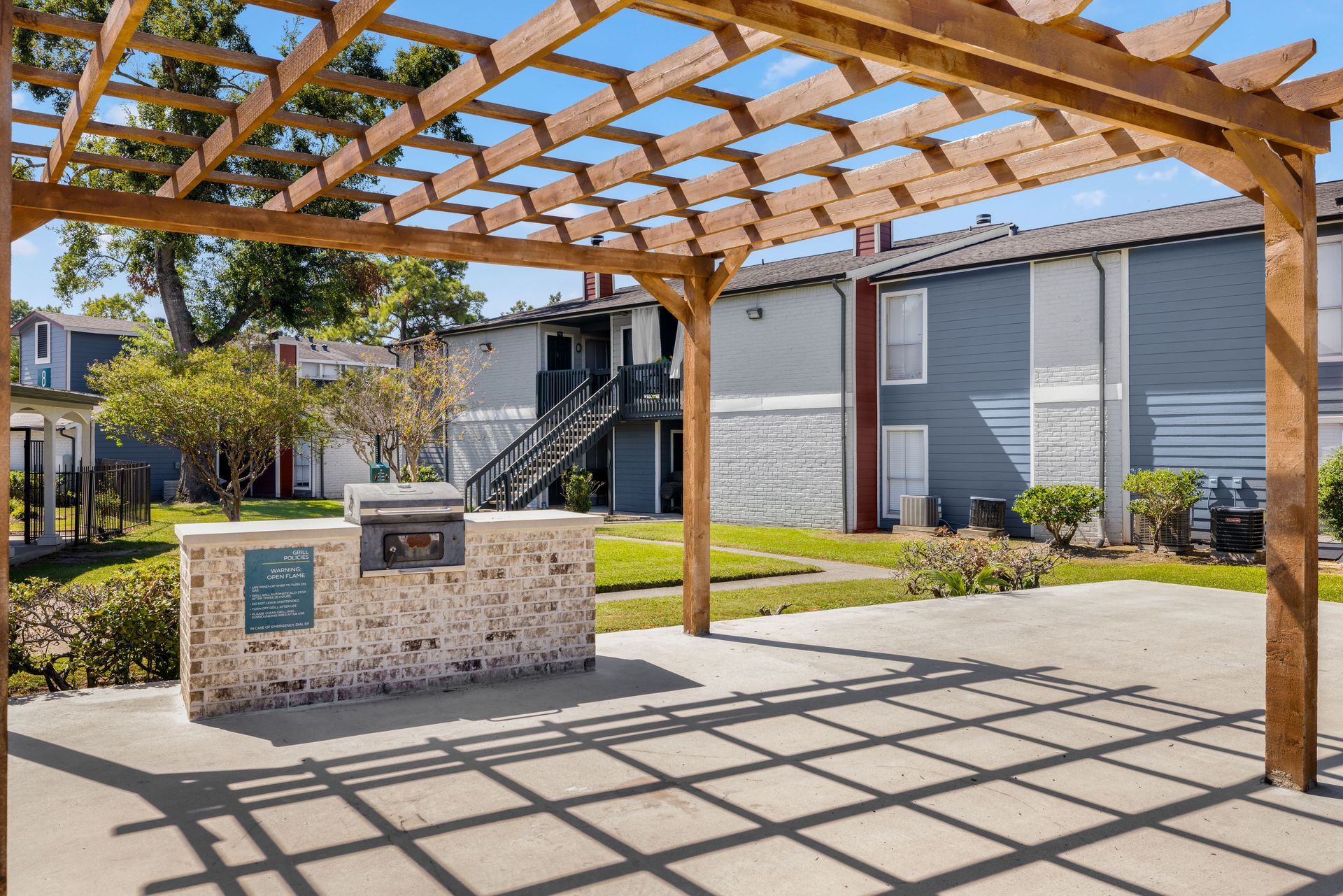 There is a grill under a pergola in front of a building at Serena Apartment Homes.