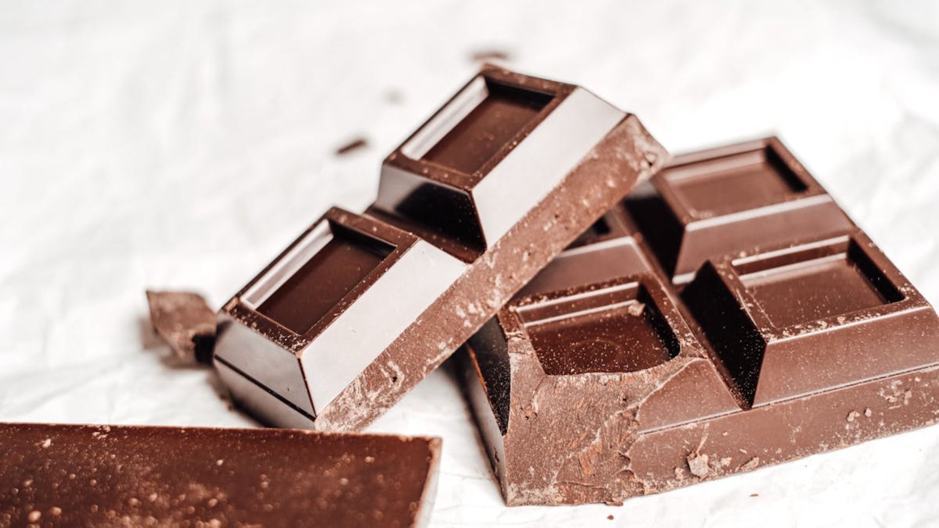 Barras de chocolate negro, rotas en pedazos, sobre una superficie blanca y arrugada.