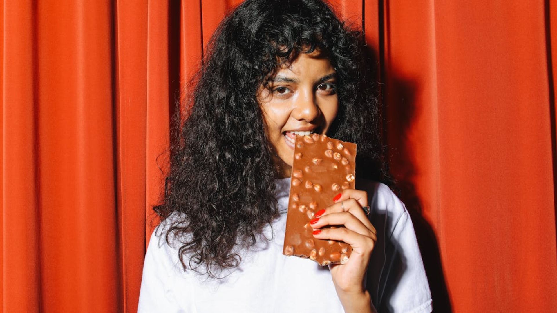 Mujer con cabello rizado, mordiendo una gran barra de chocolate, fondo de cortina roja.