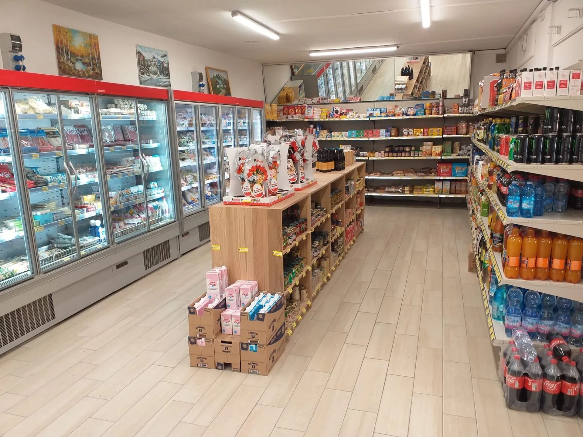 Un piccolo negozio di alimentari con vetrine refrigerate, una scaffalatura centrale in legno e corsie rifornite di merci.