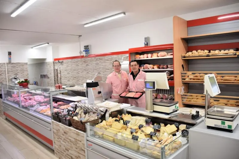 Due macellai in uniforme rosa stanno dietro un banco gastronomia colmo di salumi e formaggi all'interno di un negozio ben illuminato.