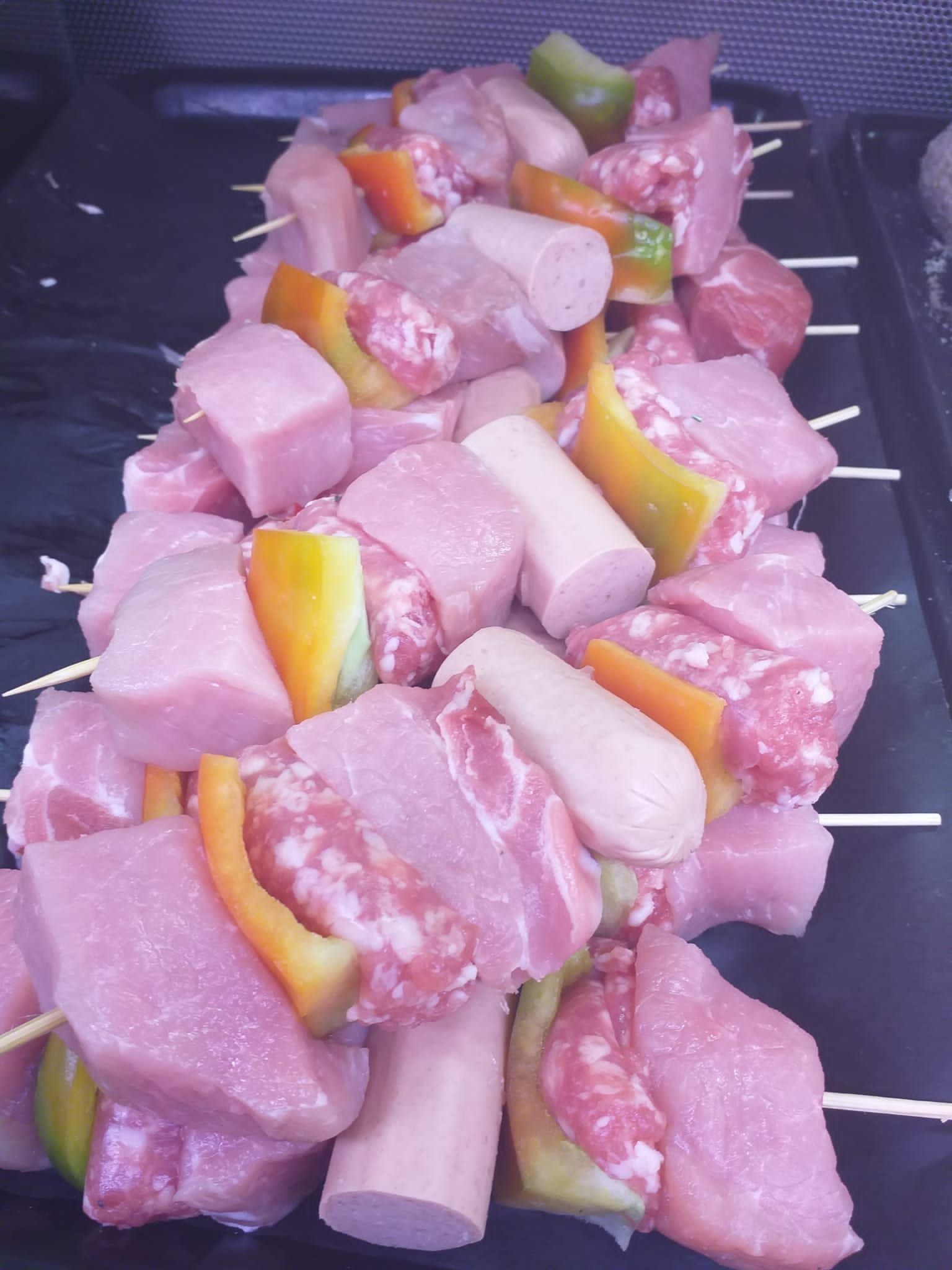 Spiedini di cubetti di maiale crudo, fette di salsiccia e pezzi di peperoni gialli e verdi su un vassoio scuro.