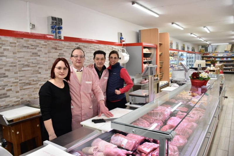 Quattro persone posano dietro una vetrina per la carne in una macelleria ben illuminata con pareti rivestite in pietra.