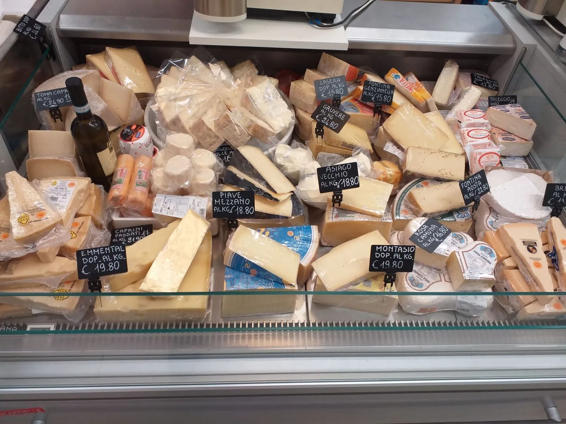 Una vetrina refrigerata piena di formaggi assortiti, disposti a spicchi e a blocchi con etichette dei prezzi.