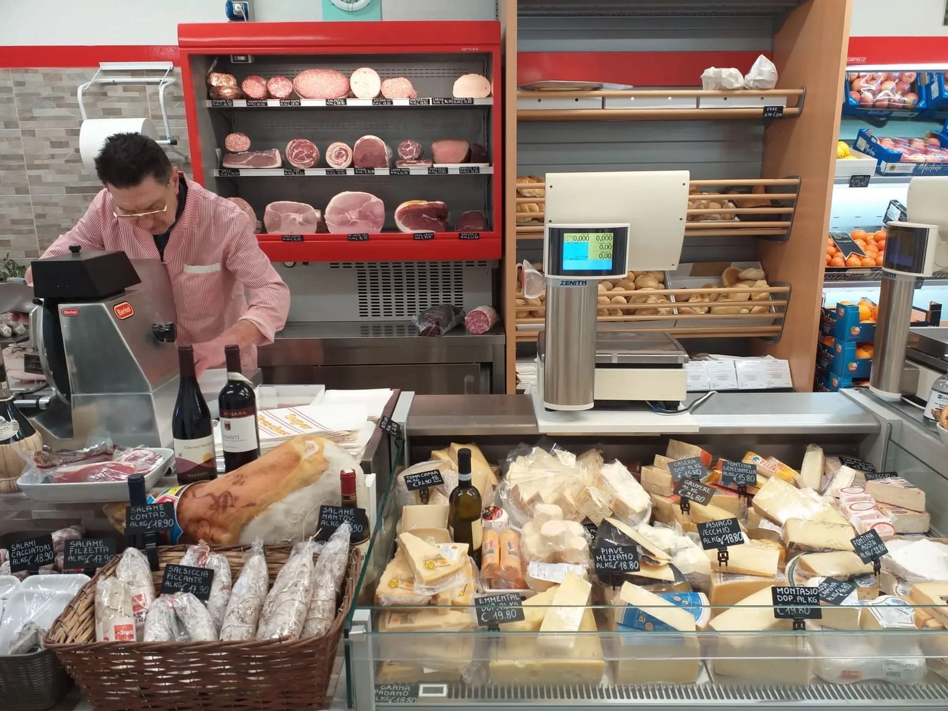 Un commesso con una camicia a quadri rossi prepara del cibo dietro un banco di gastronomia pieno di formaggi, salumi e bottiglie di vino di vario tipo.